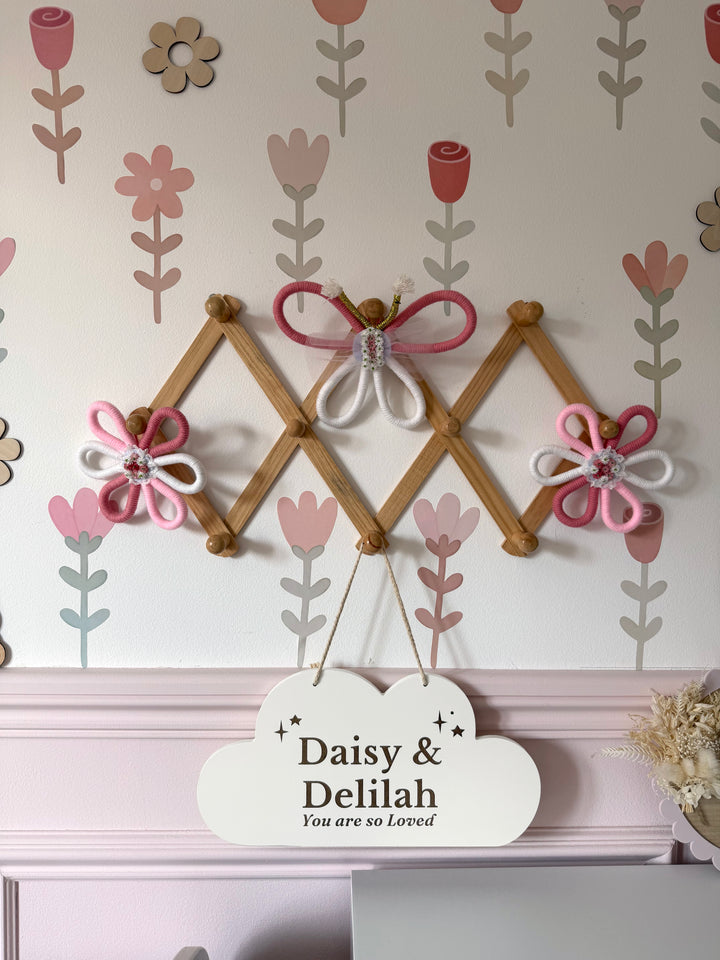 Decorative wall hanging with wooden frame, flowers, and a sign reading 'Daisy & Delilah You are so loved' against a floral wallpaper.