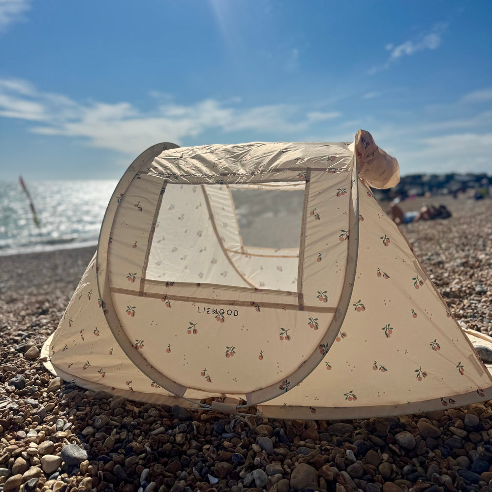 A beige pop-up beach tent with a small animal print pattern set up on a pebble beach under a bright blue sky with sun rays.