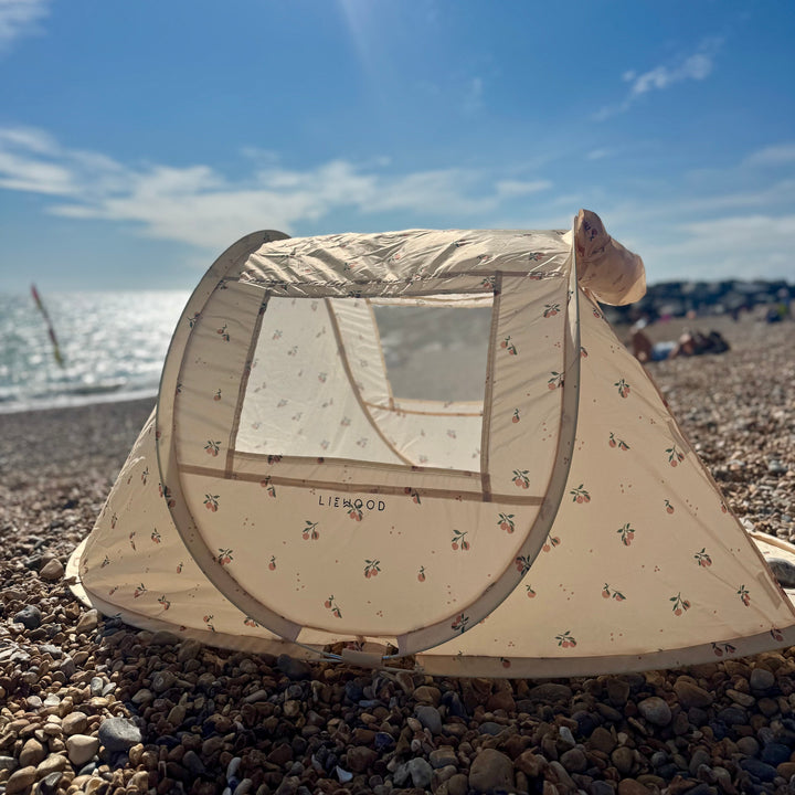 A beige pop-up beach tent with a small animal print pattern set up on a pebble beach under a bright blue sky with sun rays.