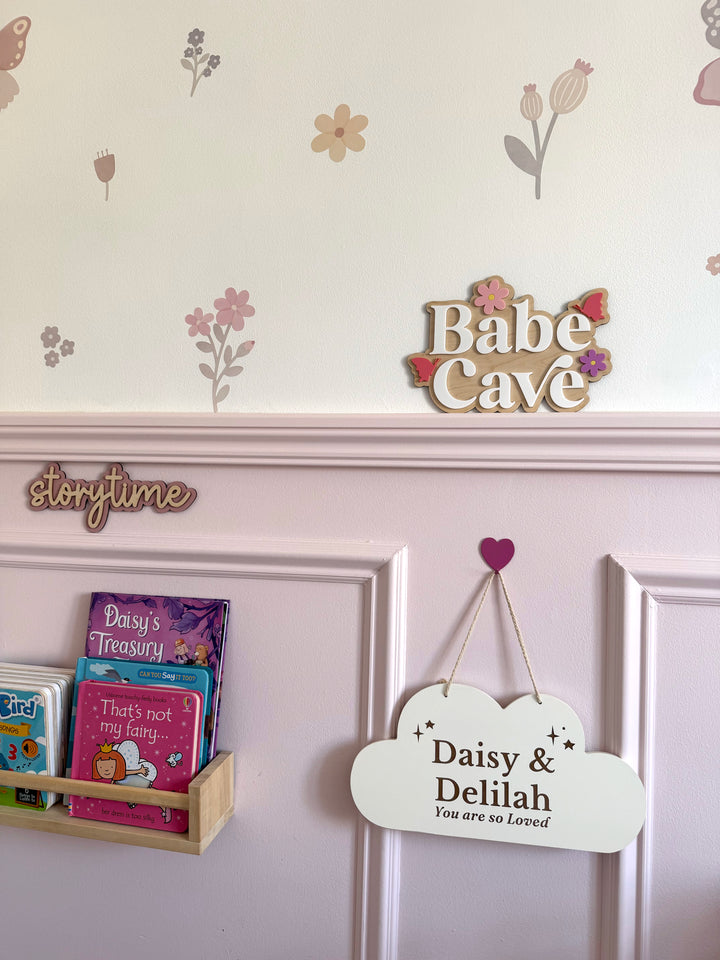 Children's room decor with 'Babe Cave' sign, books, and personalized plaque on a floral wallpapered wall.
